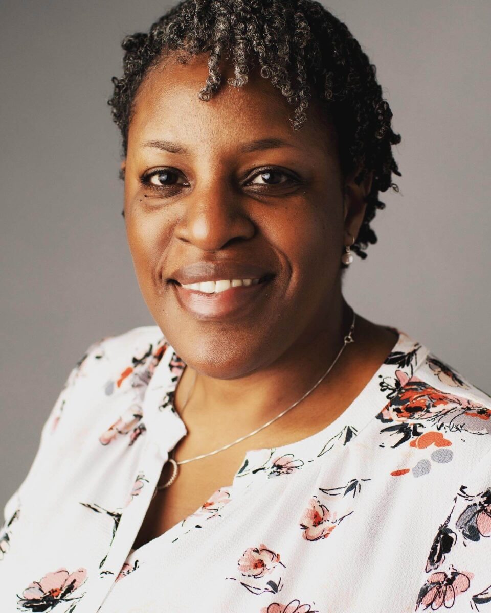 Professional portrait of a woman with short natural curls, wearing a white floral blouse and smiling in front of a neutral background.