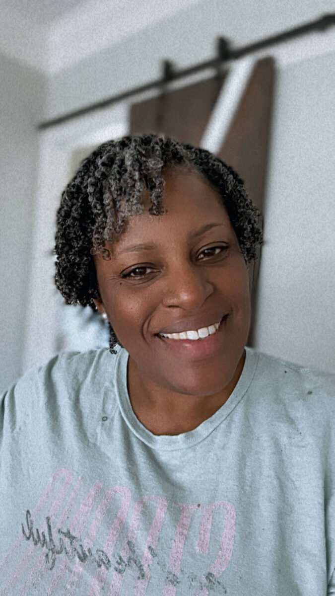 Woman with short, defined curls wearing a mint green T-shirt and smiling warmly indoors